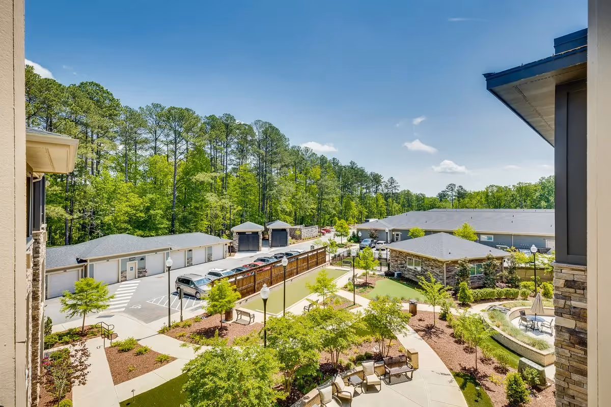 View over a landscaped courtyard with walkways, seating areas, a long putting/bocce green, parking garages and surrounding buildings backed by trees.