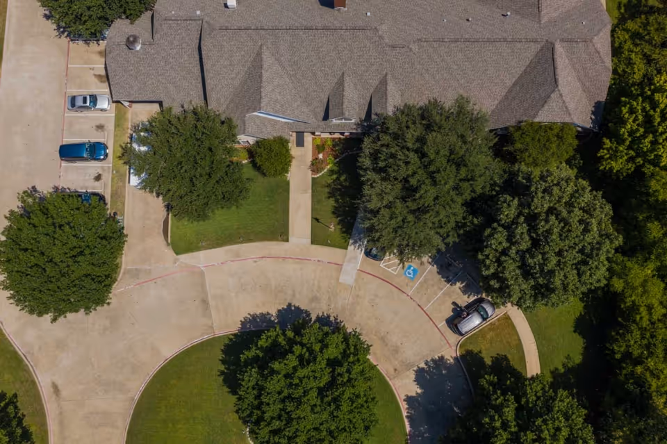Aerial view of Deer Creek Senior Living facility showing the building roof, surrounding trees, parking spaces including a handicapped spot, and a circular driveway with green lawns and sidewalks.