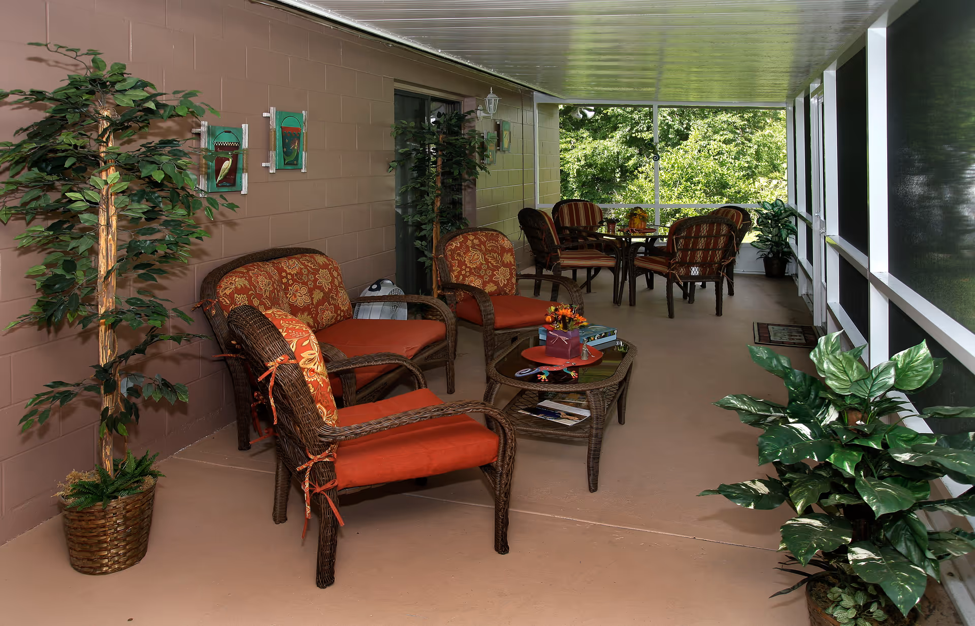 Screened outdoor seating area with wicker chairs and tables, red cushions, potted plants, and a view of greenery.