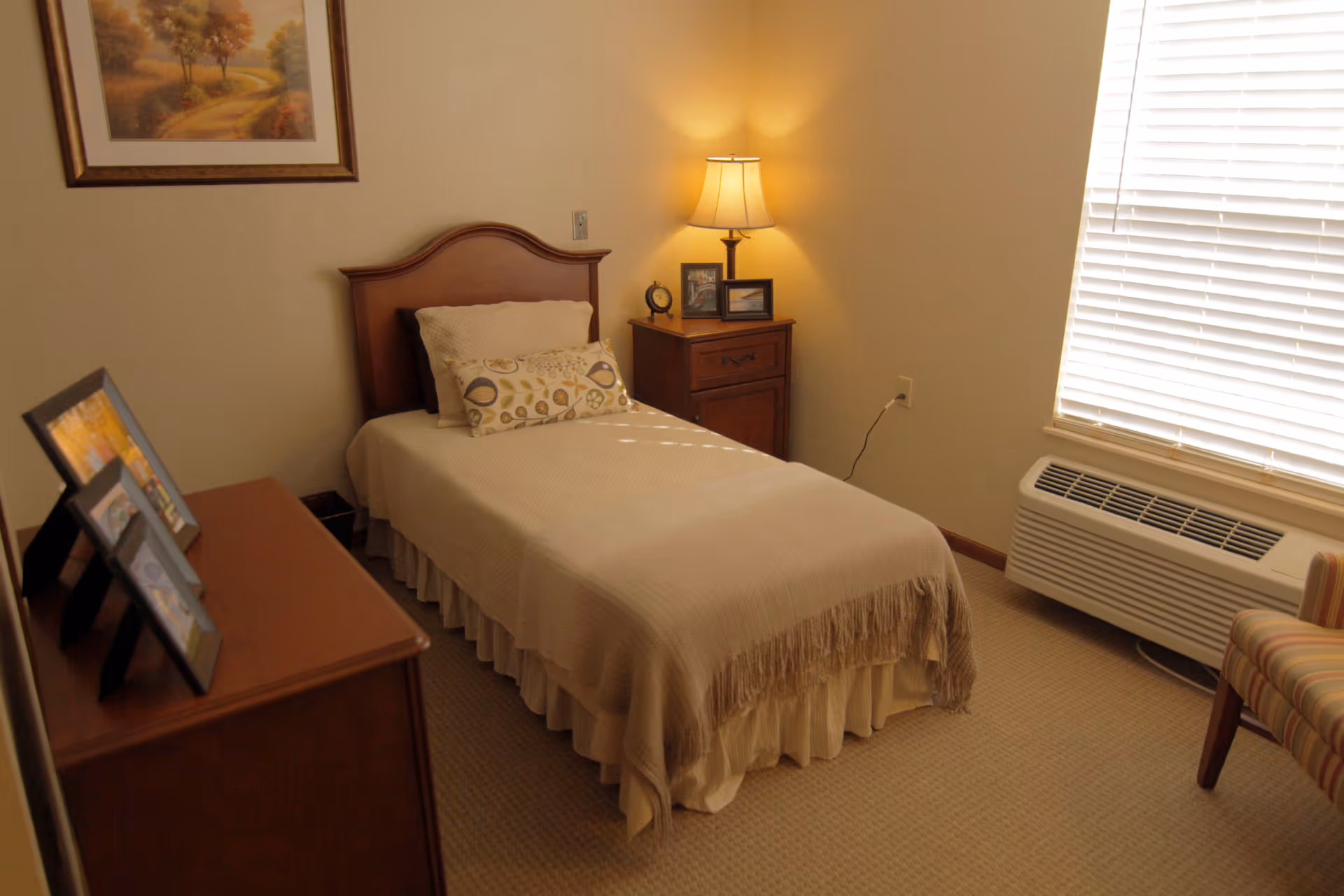 Small furnished bedroom with a single bed, nightstand and lamp, dresser with framed photos, and a window with blinds.