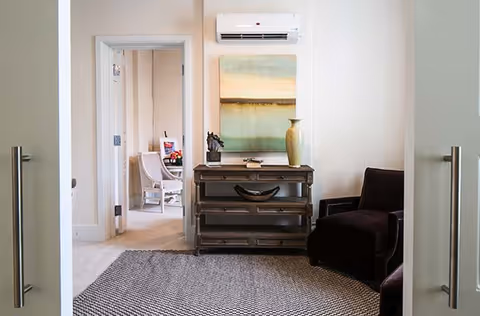 Small living area seen through double doors with a console table, vase and painting under a wall-mounted air conditioner, a dark armchair and patterned rug.
