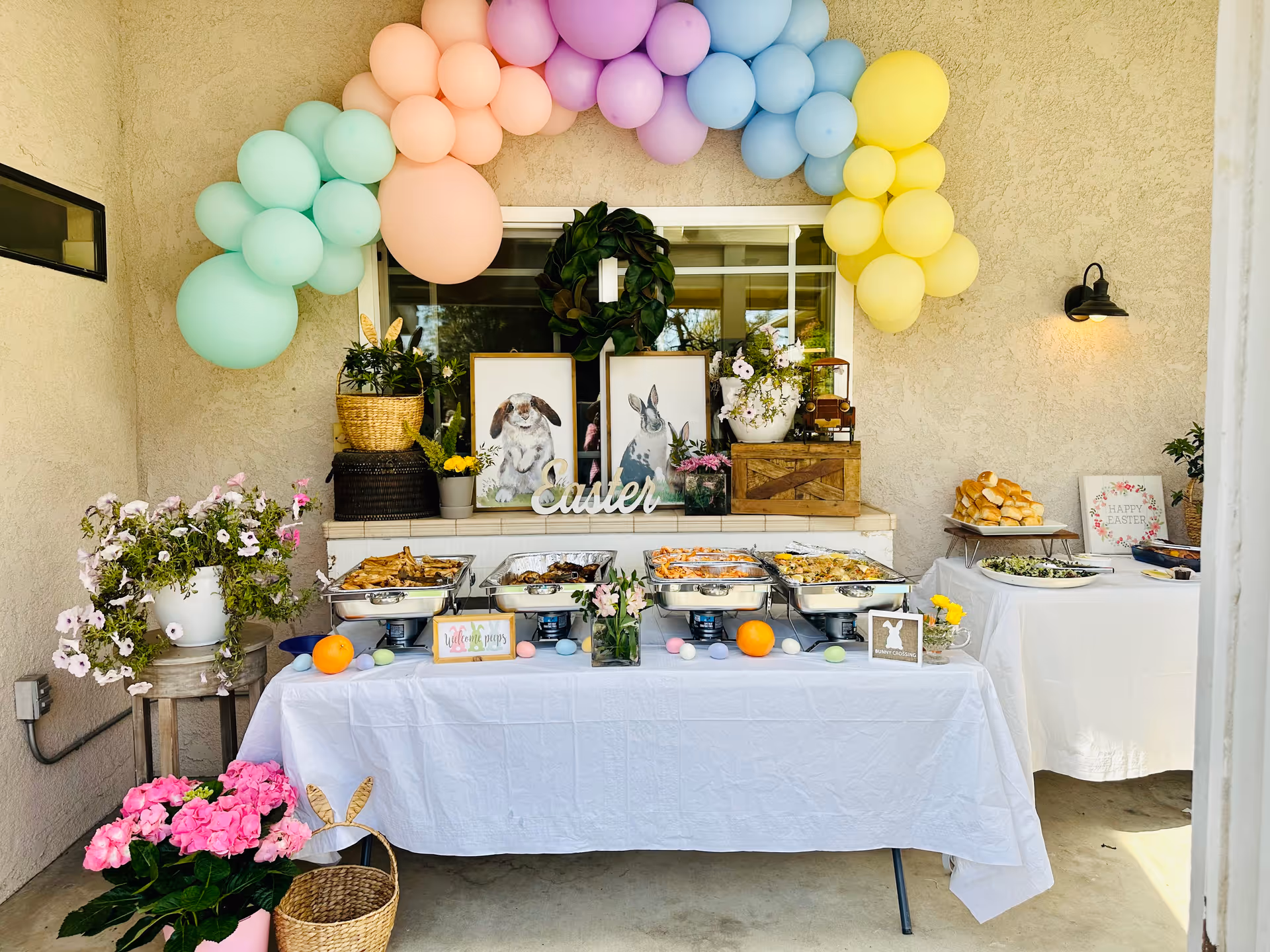 Outdoor buffet setup for an Easter celebration with pastel-colored balloon arch overhead. The buffet table is covered with a white tablecloth and holds several chafing dishes with food, decorated with Easter-themed signs and pastel-colored eggs. Behind the table are framed pictures of rabbits, a green wreath, and flower arrangements. Another table to the right holds bread rolls and salad. Potted flowers and plants decorate the area.