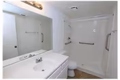 A clean, white bathroom featuring a sink with a countertop and cabinet, a large mirror above the sink, a toilet, and a bathtub with a showerhead and grab bars on the walls for safety.