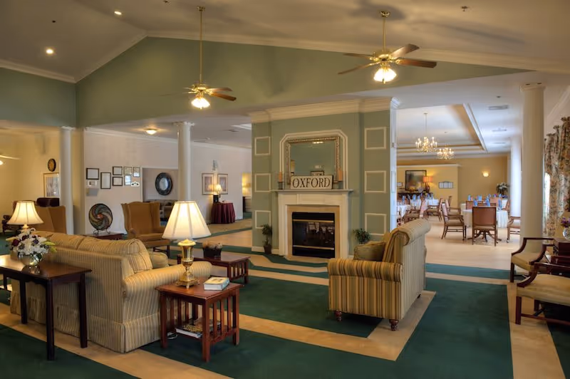 A spacious and well-lit common area in a senior living facility featuring comfortable striped sofas, wooden side tables with lamps, a fireplace with a mirror and a sign that says 'OXFORD', ceiling fans, and a dining area with tables and chairs in the background.