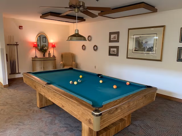 A room with a wooden pool table with a green felt surface and several billiard balls scattered on it. The room has beige walls adorned with framed pictures and decorative plates. There is a wooden cabinet with a mirror and two red lamps on top, and a beige armchair in the corner. A ceiling fan with a light fixture hangs above the pool table.