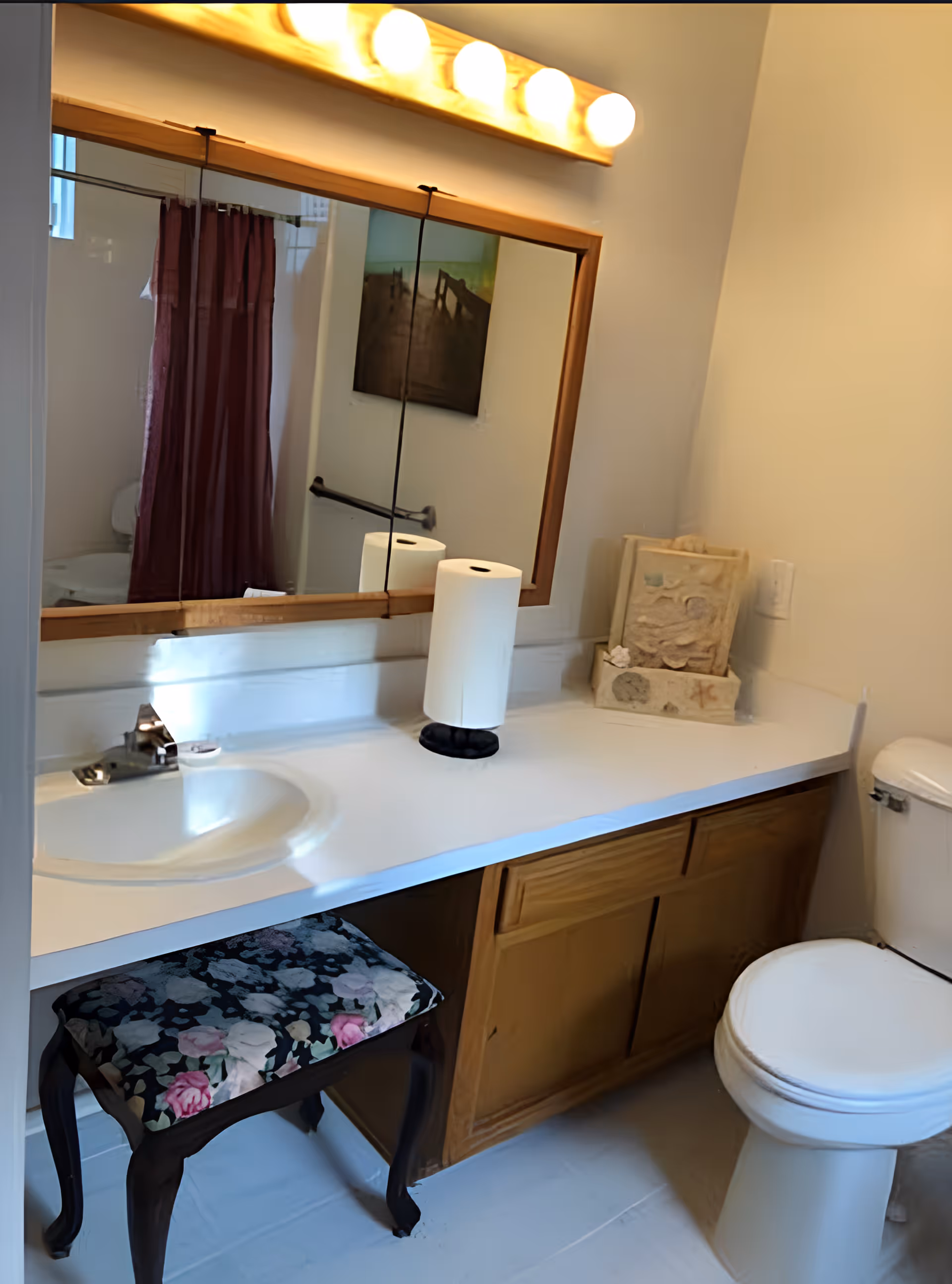 Bathroom with a white sink and countertop, wooden cabinet below, a toilet, and a large mirror above the sink. A floral cushioned stool is placed under the counter. There is a paper towel roll on the countertop and a decorative item next to it. A shower curtain and a framed picture are visible in the mirror reflection.
