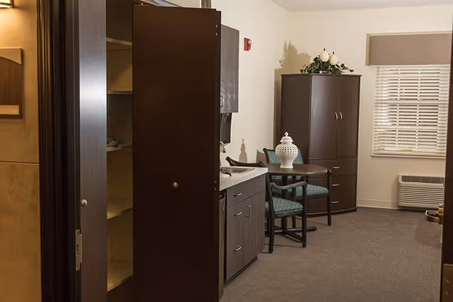 Interior view of a senior living facility room with dark wood cabinets, a small round table with two chairs, a decorative white vase on the table, a window with blinds, and a wall-mounted air conditioning unit below the window.