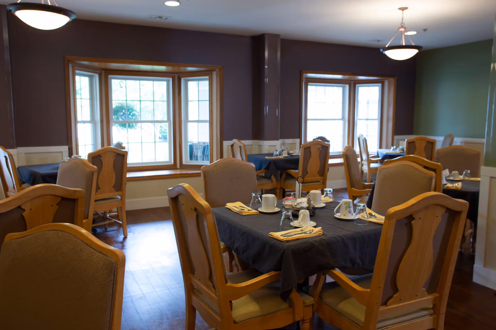 Dining room with several wooden tables and chairs arranged neatly. Tables are covered with dark tablecloths and set with cups, glasses, napkins, and utensils. Large windows with wooden frames allow natural light to brighten the room.