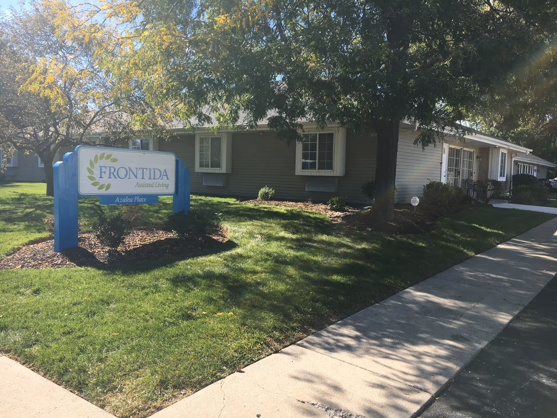 Exterior view of Frontida Assisted Living: Azalea Place building with a blue and white sign in front on a grassy area. The building is single-story with beige siding, white window frames, and surrounded by trees and shrubs. A sidewalk runs alongside the building.