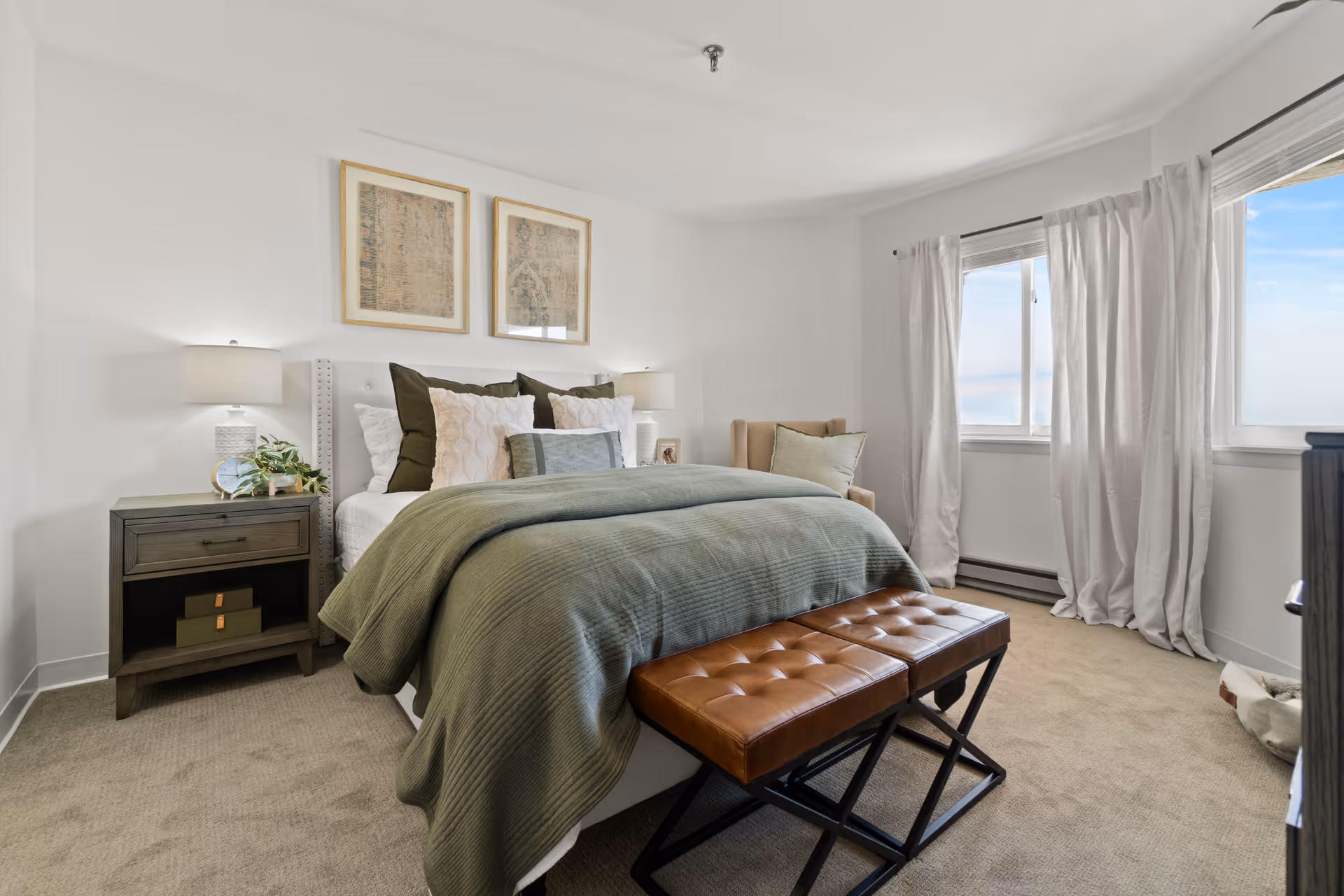 Bright, neutral bedroom with a neatly made bed dressed in green bedding, bedside tables with lamps, a leather bench at the foot of the bed, and windows with curtains.