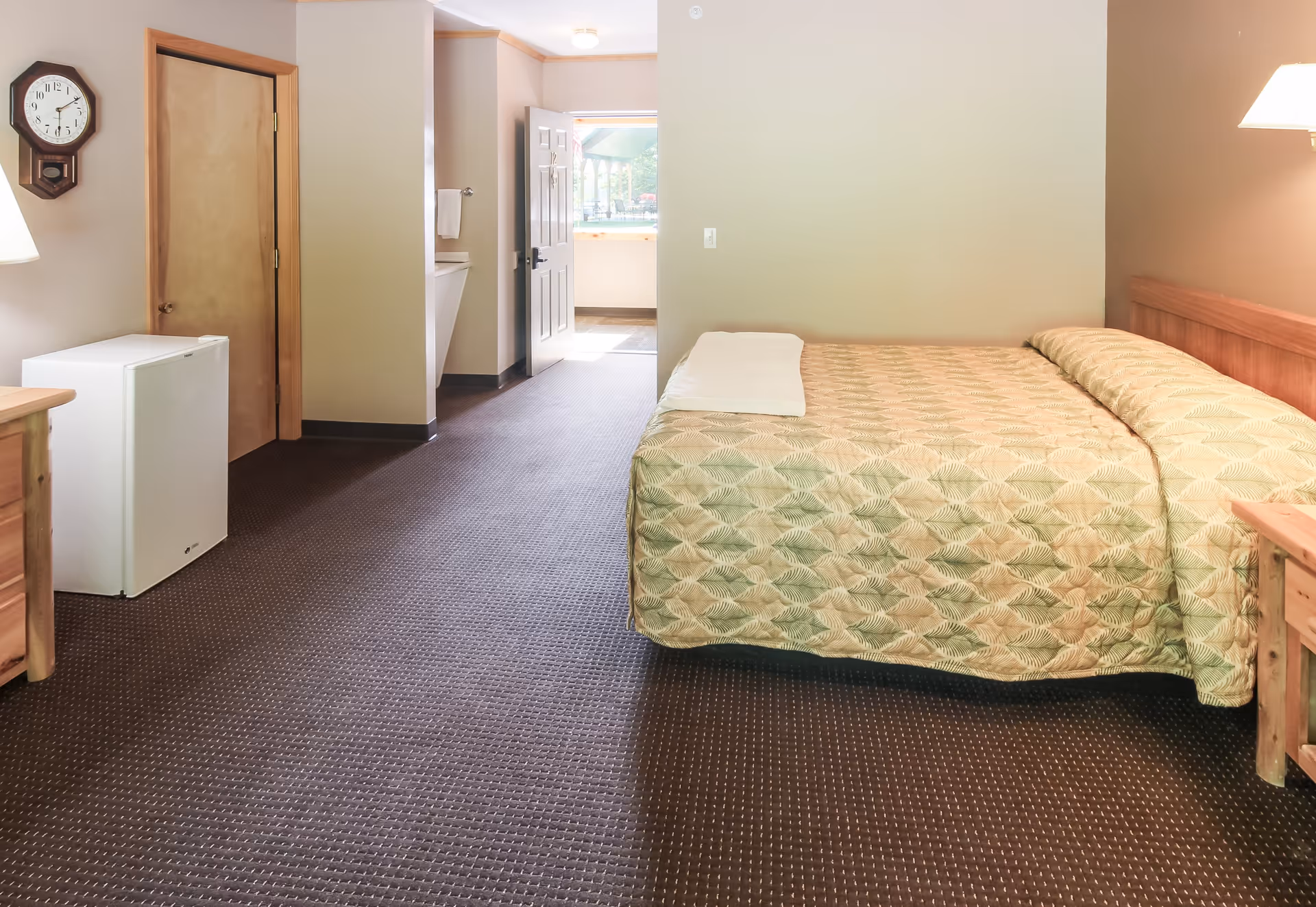 A simple bedroom in an assisted living facility featuring a bed with a patterned bedspread, a wooden nightstand with a lamp, a small white refrigerator, a wall clock, and an open door leading to a hallway with a sink and towel rack.