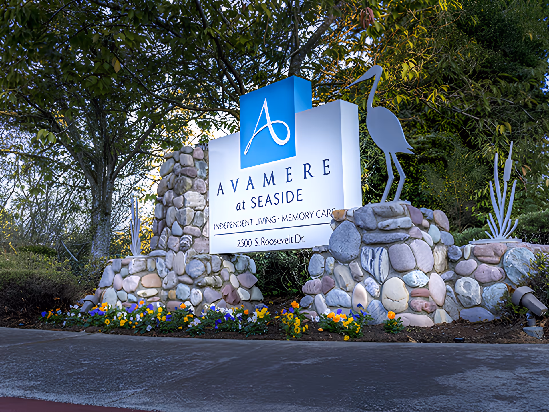Stone and rock base sign for Avamere at Seaside senior living facility with a blue logo and white text, surrounded by flowers and greenery, with metal sculptures of a bird and plants on either side.