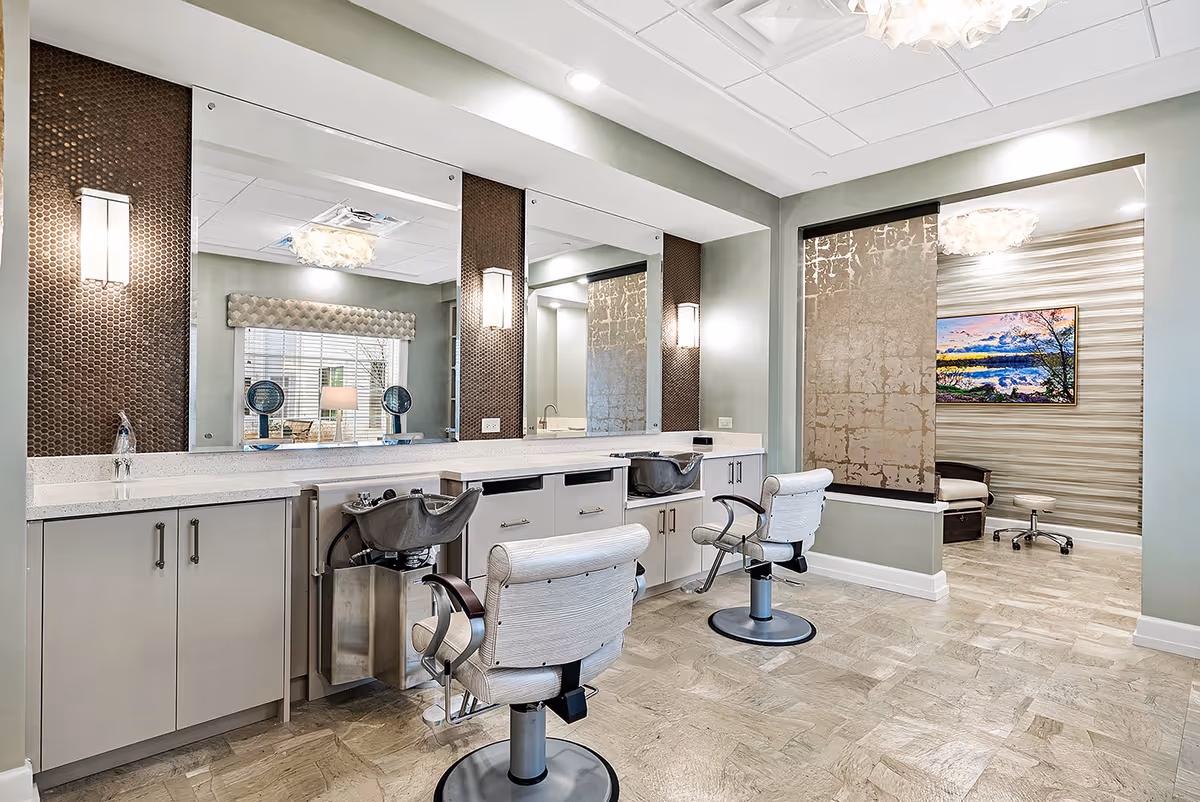 Interior view of a salon area in a senior living facility with two salon chairs in front of large mirrors and wash basins. The room features soft green walls, modern lighting fixtures, and a separate seating area with a chair and a colorful landscape painting on the wall.