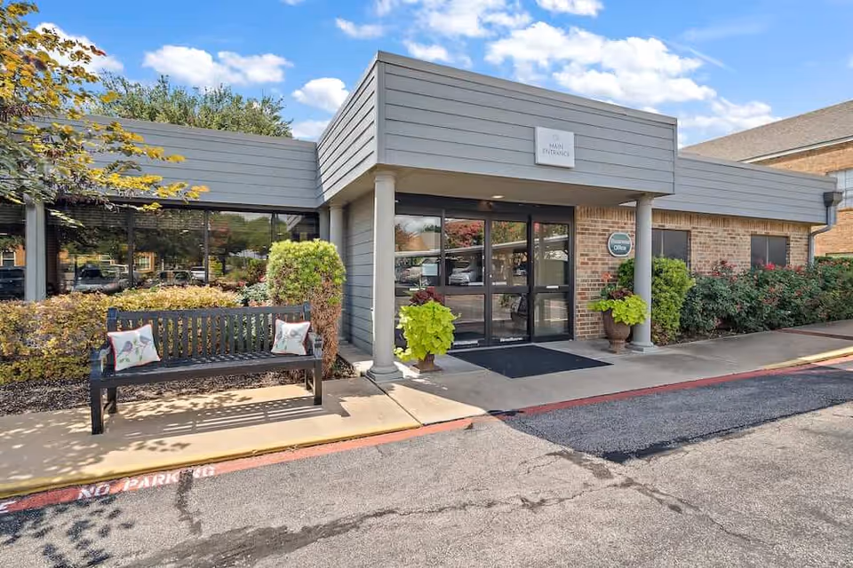 Exterior view of the main entrance of a single-story building with gray siding and brick walls. There is a black bench with two decorative pillows on the left side, surrounded by bushes and plants. Two large potted plants flank the entrance door, which has glass panels. The sky is blue with some clouds.