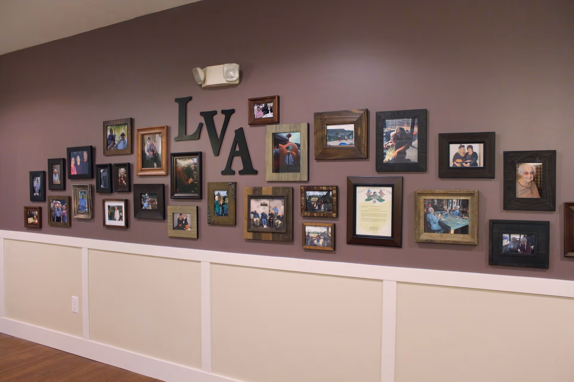 A wall decorated with multiple framed photographs and the letters LVA arranged in the center. The wall is painted a muted purple color above white wainscoting, and the floor is wooden. The photos depict various people and moments, creating a warm and personal atmosphere.