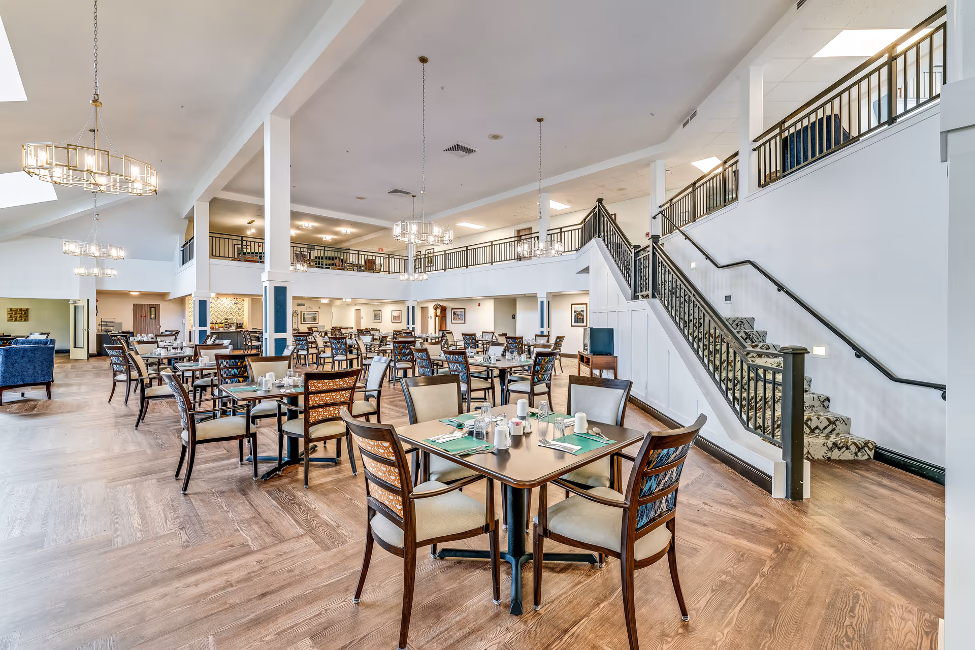 Spacious dining room with multiple set tables and chairs, chandeliers, and a staircase leading to an upper level.
