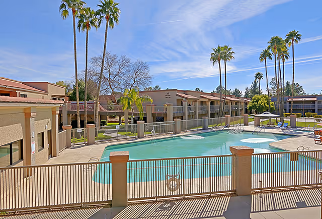 Outdoor swimming pool area at Pueblo Norte Senior Living with a fenced pool, lounge chairs, umbrellas, palm trees, and two-story residential buildings in the background under a clear blue sky.