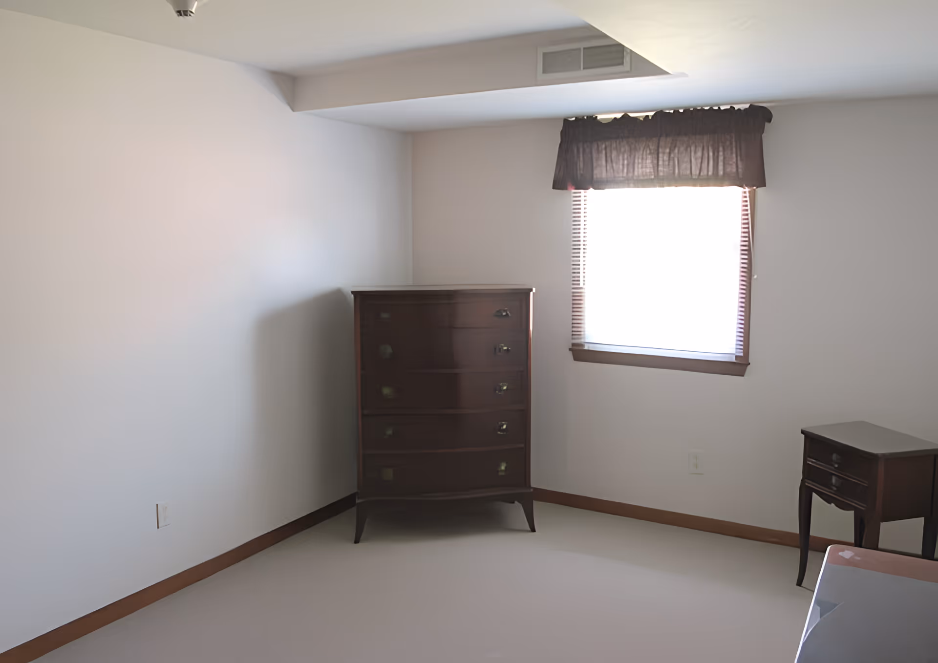 Sparsely furnished bedroom with a tall wooden dresser, small side table, and a window with a valance.