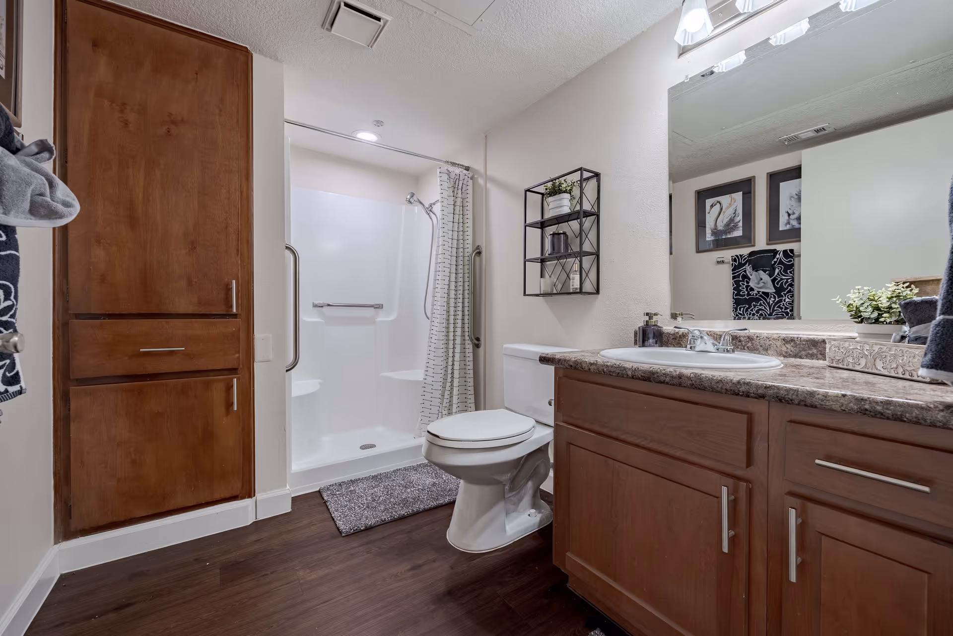 A clean and modern bathroom featuring a white toilet, a shower with a white curtain and built-in seats, a wooden cabinet, and a countertop with a sink. The walls are light-colored, and there is a large mirror above the sink. A small black metal shelf with decorative items is mounted on the wall above the toilet. The floor is dark wood, and there are framed pictures and towels visible in the reflection of the mirror.