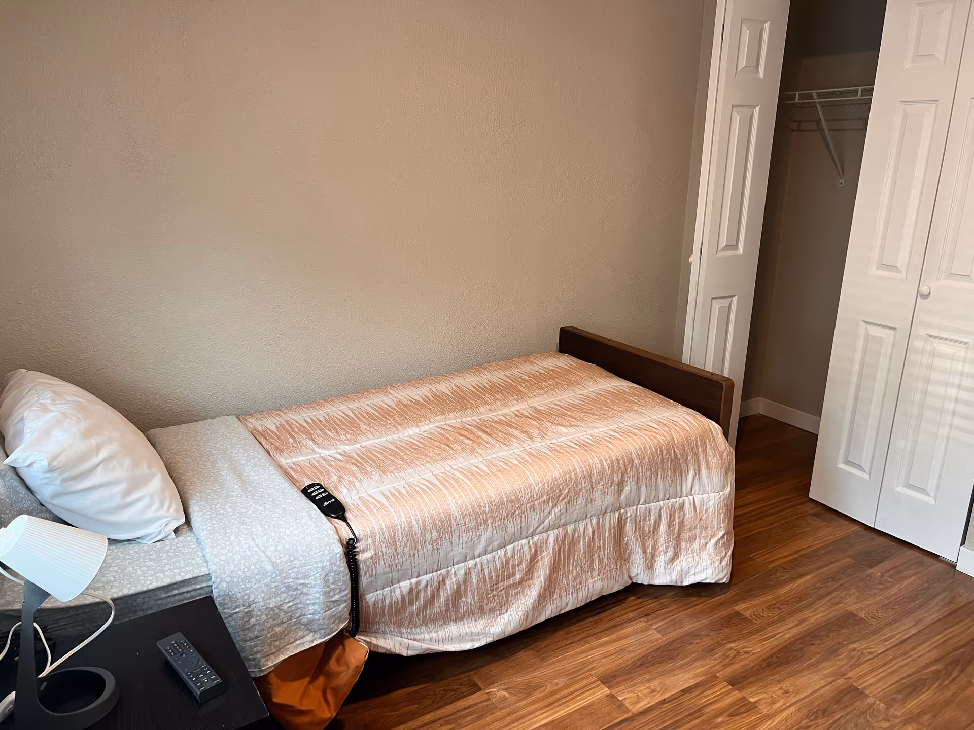 A small bedroom with a single bed covered with a peach-colored bedspread and a white pillow. Next to the bed is a black nightstand with a white lamp and a remote control. There is an open closet with white bi-fold doors and wooden flooring.