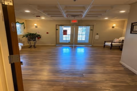 Interior view of a hallway or common area with wooden flooring, beige walls, a double glass door at the end, a bench with cushions on the right side, a plant on a decorative stand on the left, and ceiling lights.