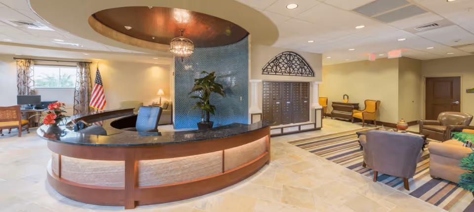 Reception area of a senior living facility with a curved wooden desk, a blue office chair, a large plant, and a decorative blue tile wall behind the desk. The room has beige tiled floors, a striped rug, several armchairs, a piano, and an American flag near a window with patterned curtains.