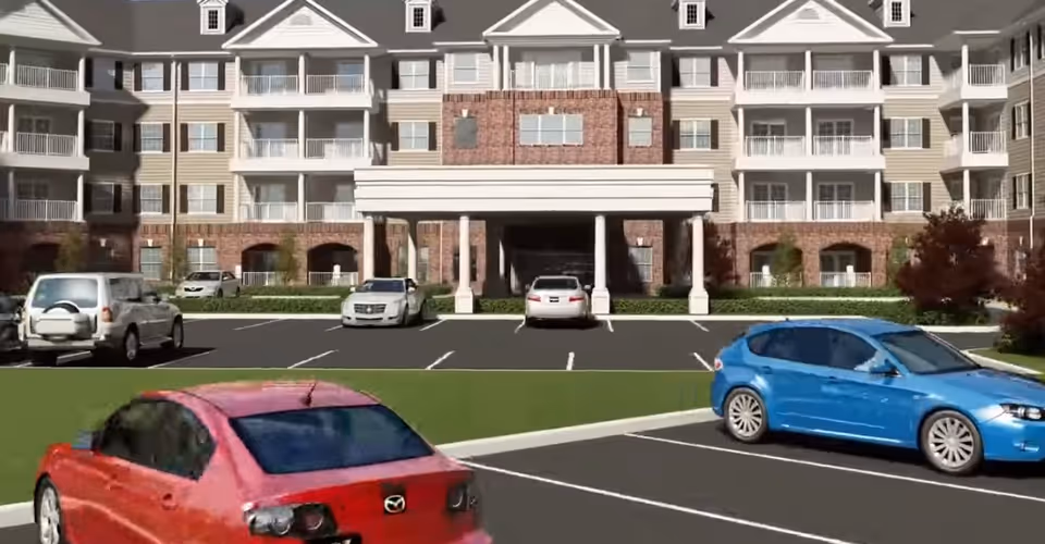 Exterior view of a multi-story senior living facility with balconies and a covered entrance. Several cars are parked in the parking lot in front of the building.
