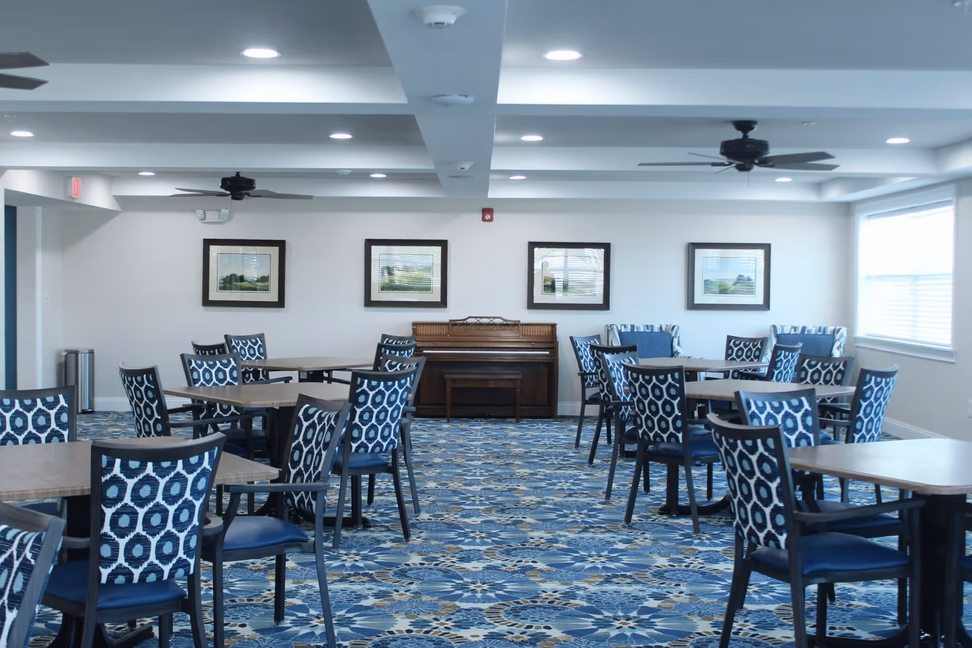 A spacious dining room with multiple tables and chairs featuring blue patterned upholstery. The room has a blue and beige patterned carpet, white walls with framed landscape paintings, ceiling fans, and large windows letting in natural light. A piano is placed against the far wall.
