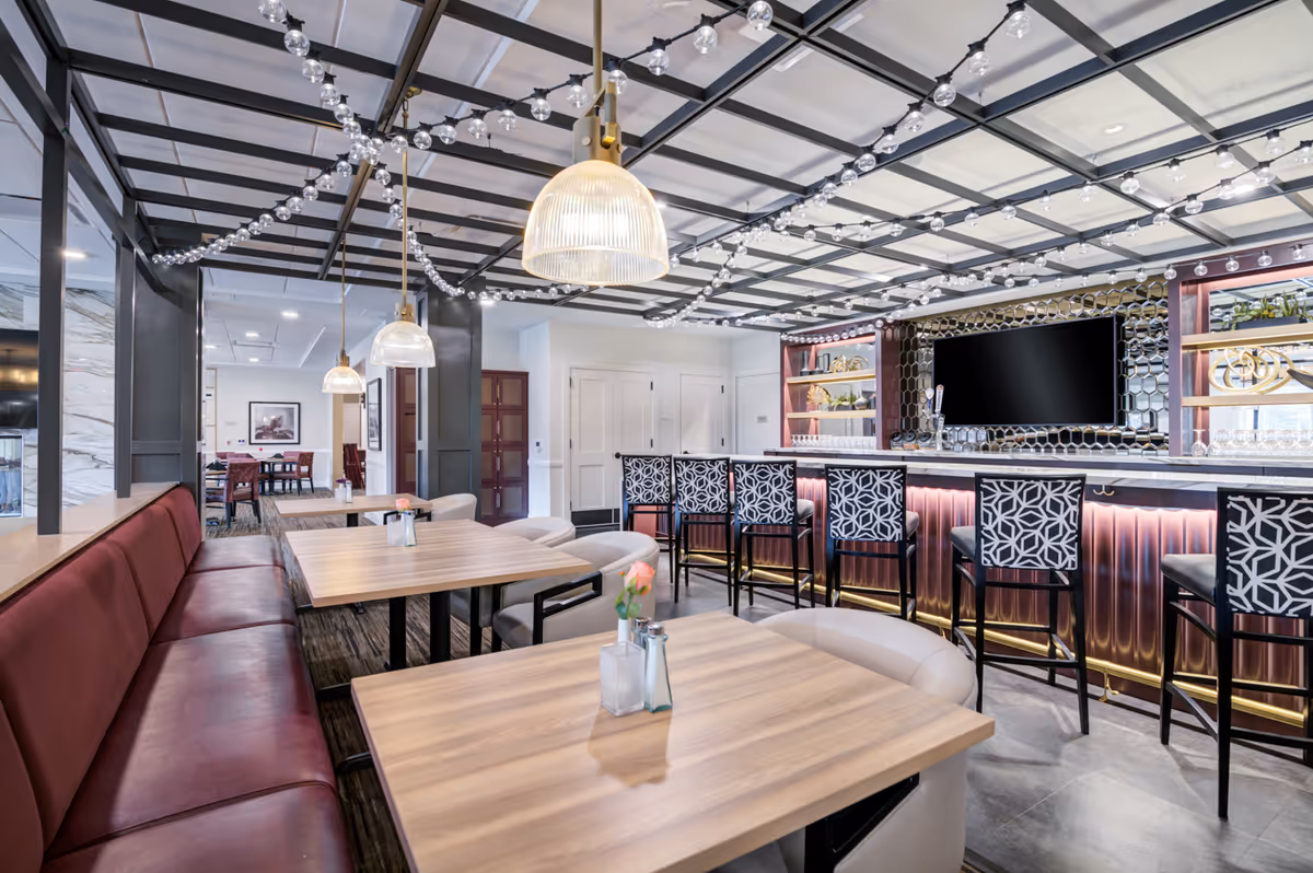 Modern dining and bar area with wooden tables, upholstered bench seating, pendant lights, and a row of patterned bar stools.