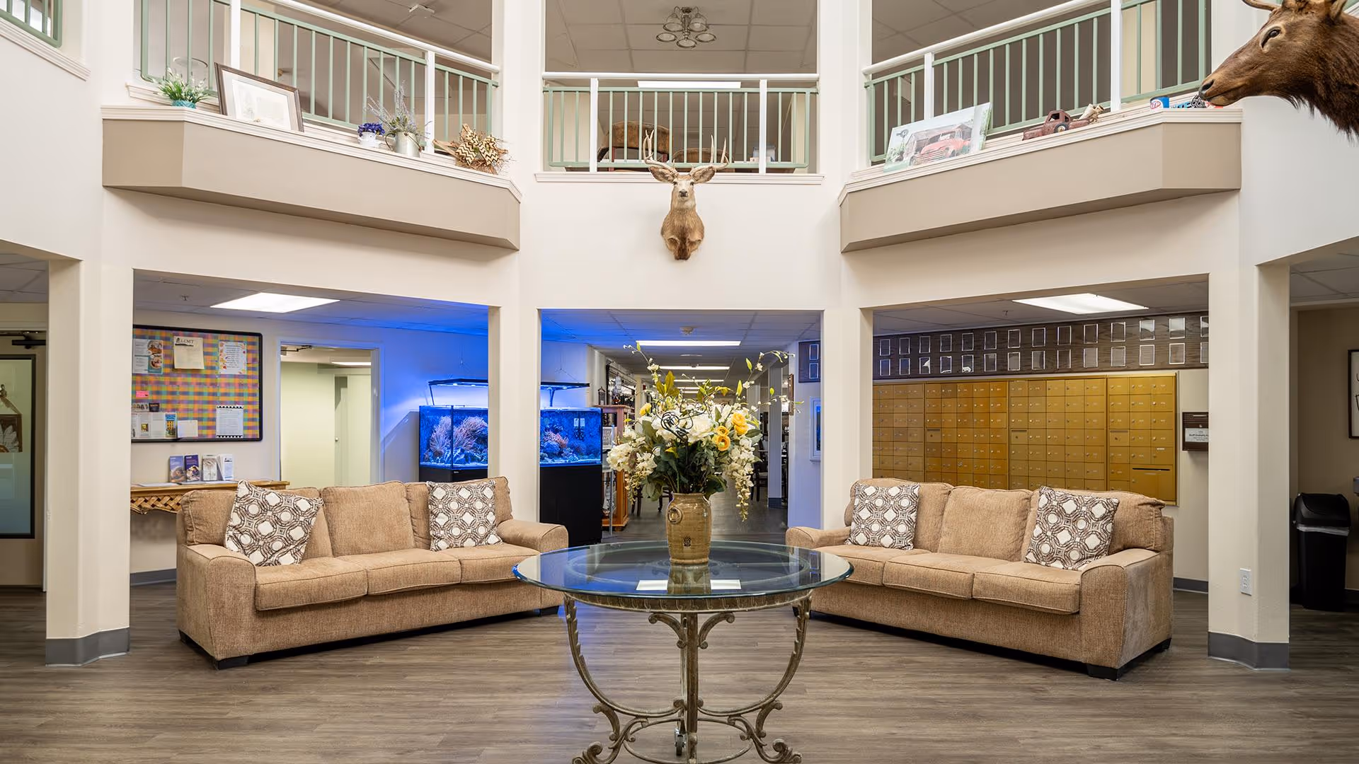 A spacious senior living facility common area with two beige sofas facing each other, a round glass table with a floral arrangement in the center, mounted deer heads on the walls, and a set of mailboxes in the background. The area has a high ceiling with a balcony railing above and fish tanks illuminated with blue light.