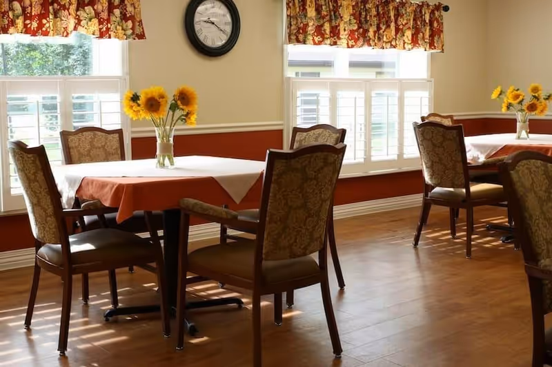 Sunlit dining room with tables covered in tablecloths, upholstered chairs, sunflower centerpieces, and windows with floral valances.