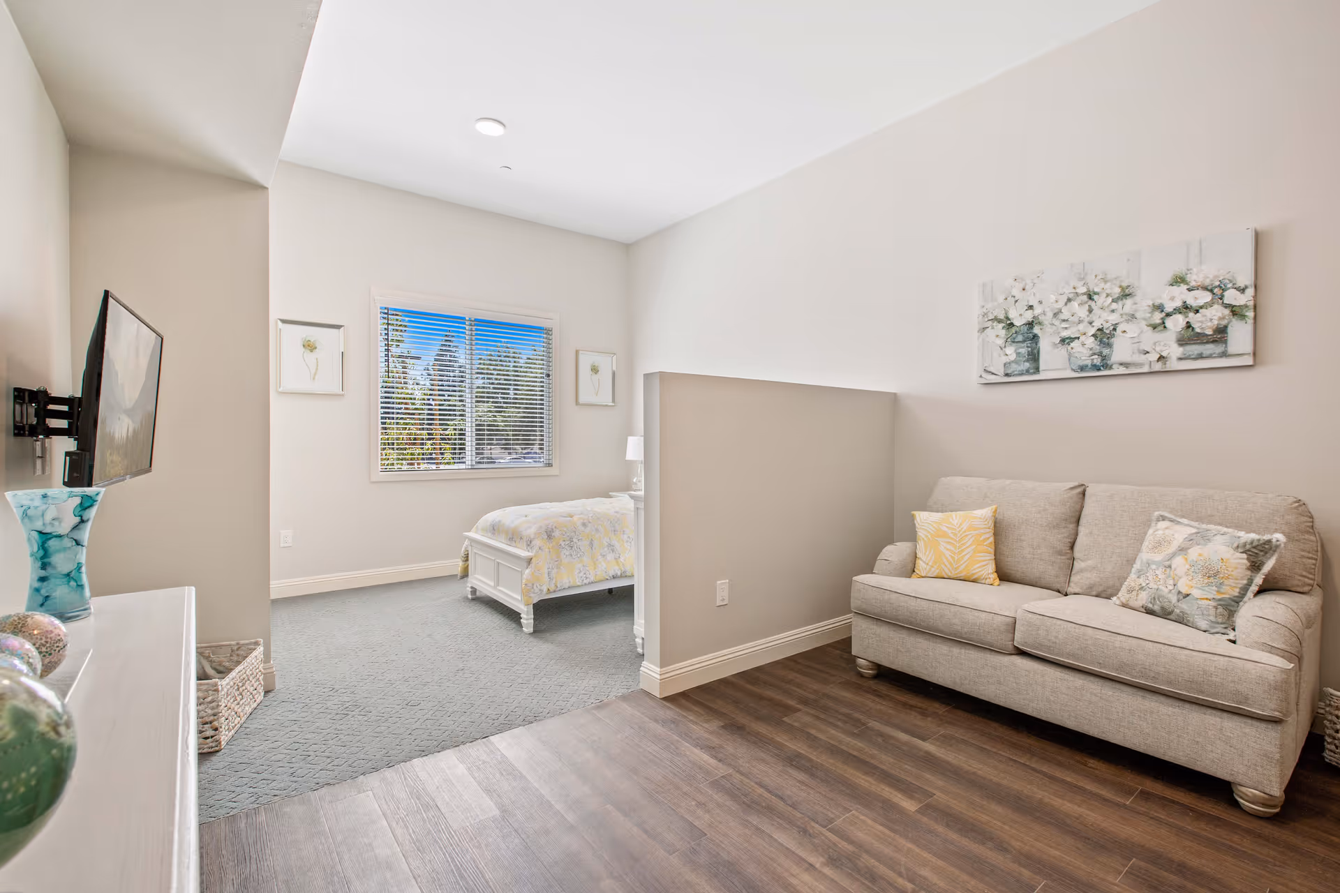 Open interior space with a beige sofa, a half-wall divider and a bed by a window under bright natural light.