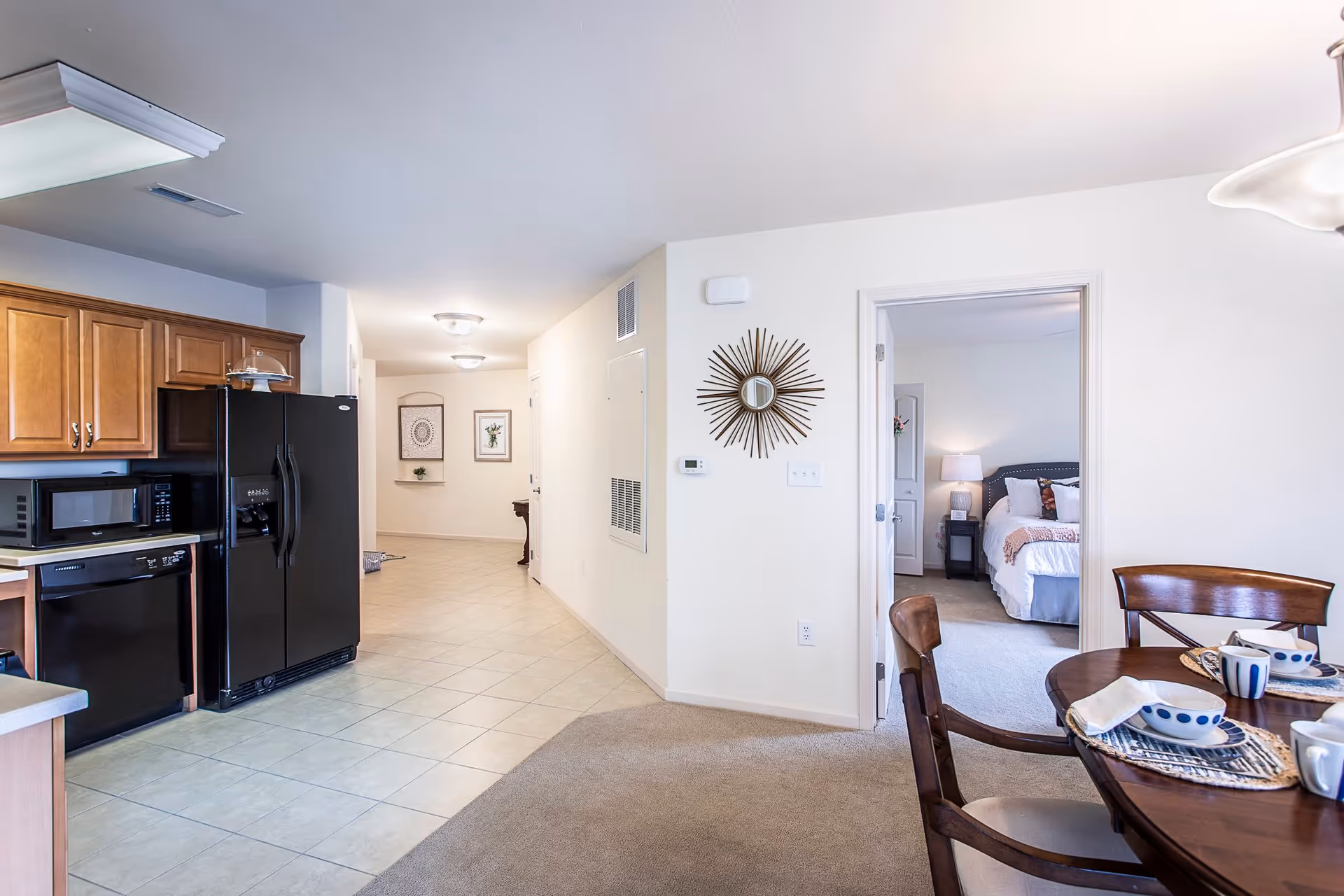 Interior view of a senior living facility showing a kitchen area with wooden cabinets, a black refrigerator, microwave, and dishwasher. Adjacent to the kitchen is a dining area with a round wooden table set with dishes and cups. A hallway leads to a bedroom with a bed, nightstand, and lamp visible through an open door. The walls are light-colored and the floor transitions from tile in the kitchen to carpet in the dining and bedroom areas.