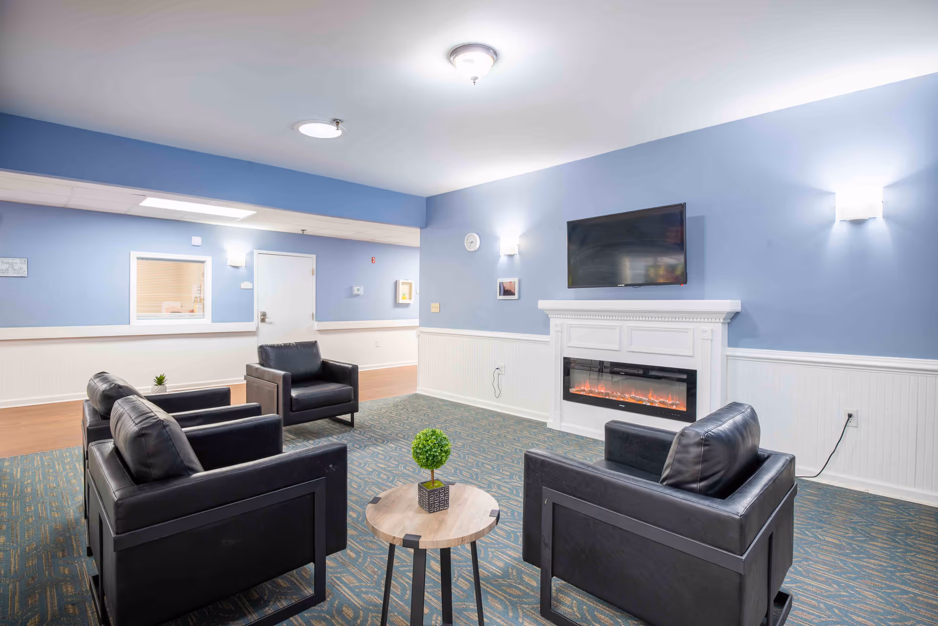 A cozy senior living common area with four black leather armchairs arranged around a small round wooden table with a small green plant. The room has blue walls with white wainscoting, a wall-mounted flat-screen TV above an electric fireplace, and soft lighting fixtures on the walls.