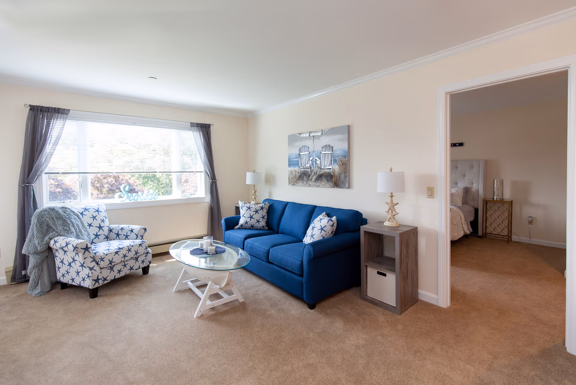 Sunlit living room featuring a blue sofa, star-patterned armchair, glass coffee table, and a doorway leading to a bedroom.