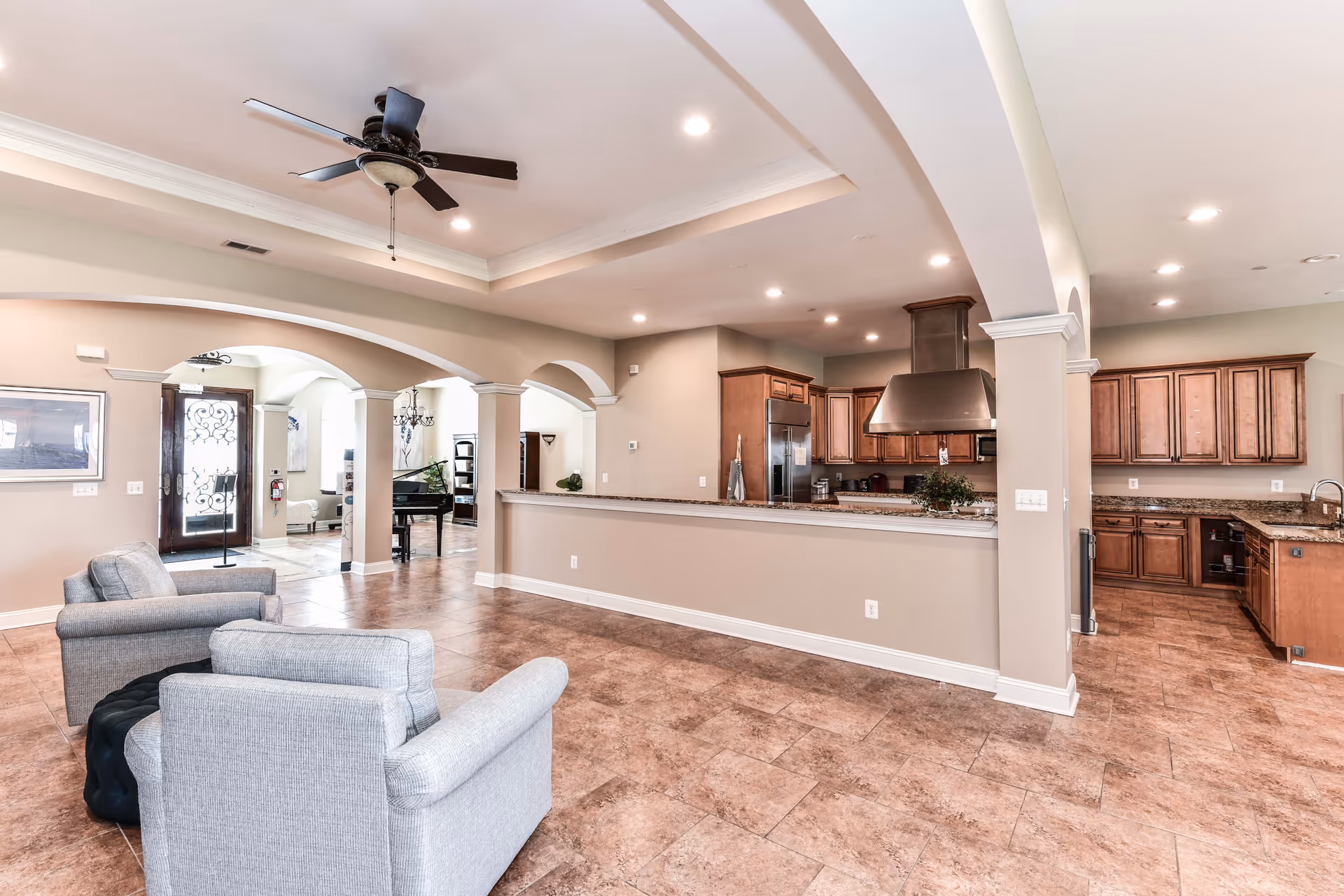 Spacious interior of a senior living facility showing a seating area with two gray armchairs, a ceiling fan, and an open kitchen with wooden cabinets and granite countertops. The room features arched doorways and tiled flooring, with a piano visible in the background near the entrance door.