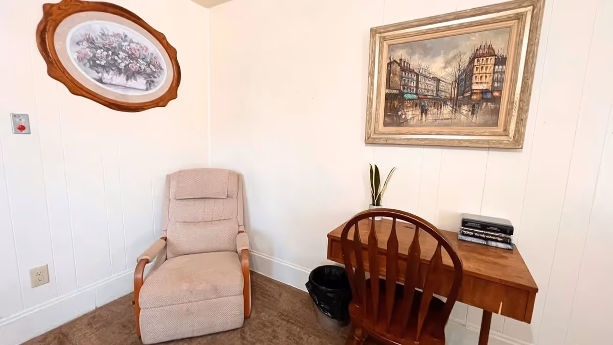 A small corner of a room with a beige upholstered armchair on the left and a wooden desk with a wooden chair on the right. On the desk are a small plant and a stack of books. The walls are white with vertical paneling, and two framed pictures hang on the walls above the chair and desk. The floor is carpeted in a brown color.