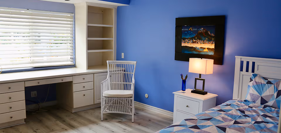 A bedroom with blue walls featuring a white wooden bed with a geometric patterned bedspread in shades of blue, white, and gray. Next to the bed is a white nightstand with a modern lamp and a blue vase with flowers. There is a white wicker chair and a built-in desk with drawers and shelves under a window with white blinds.