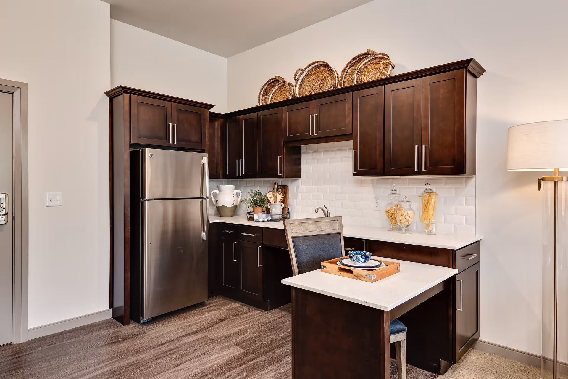 Modern kitchen area with dark wood cabinets, a stainless steel refrigerator, white countertops, and a small dining table with a chair. Decorative baskets are placed on top of the cabinets, and glass jars with pasta and snacks are on the counter. A floor lamp is visible on the right side.
