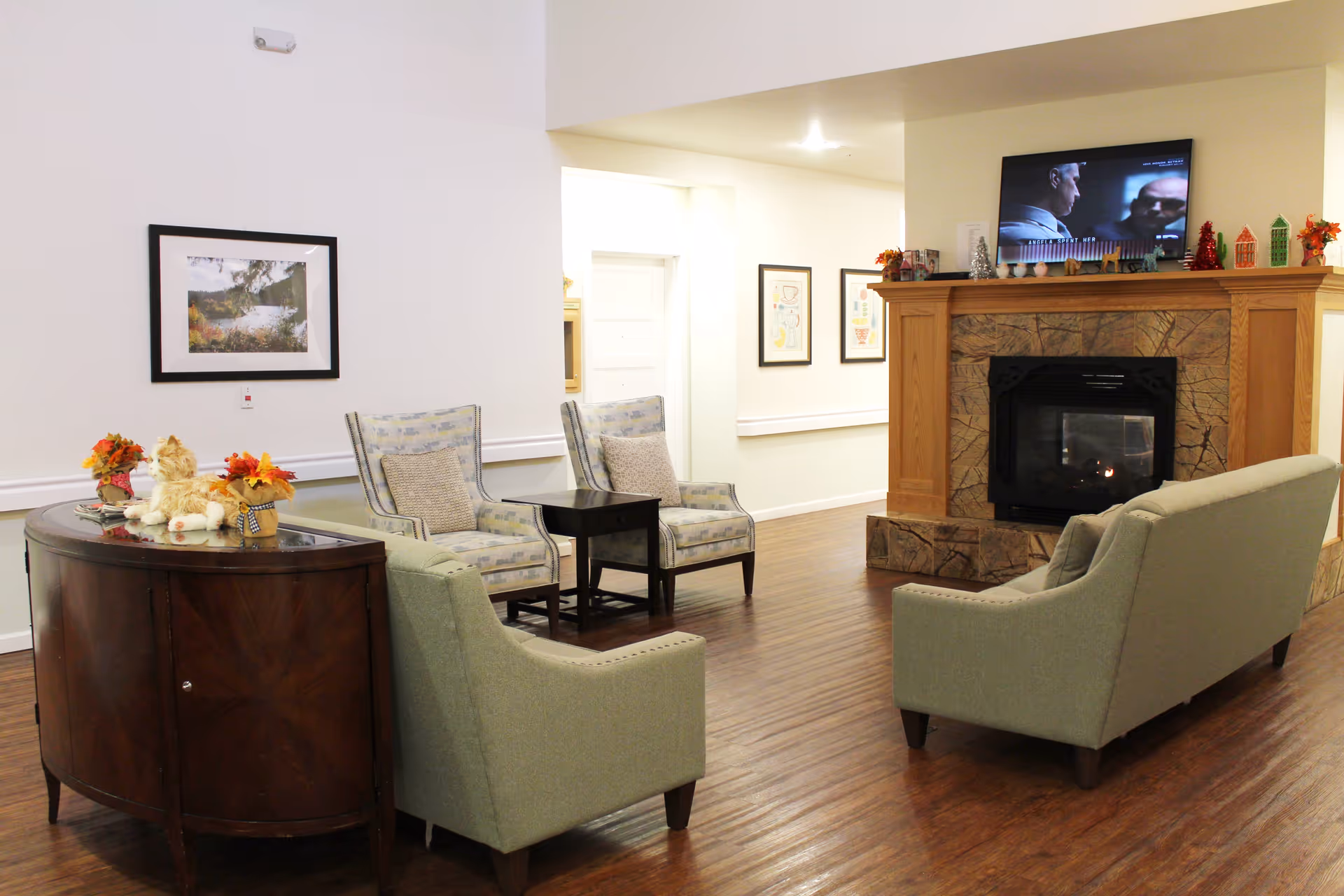 A cozy senior living facility common area with a wooden floor, two green armchairs, two patterned armchairs with cushions, a dark wooden side table, and a wooden cabinet with decorative items including a stuffed cat and small flower arrangements. A fireplace with a stone surround and a TV mounted above it is visible, along with framed artwork on the walls.