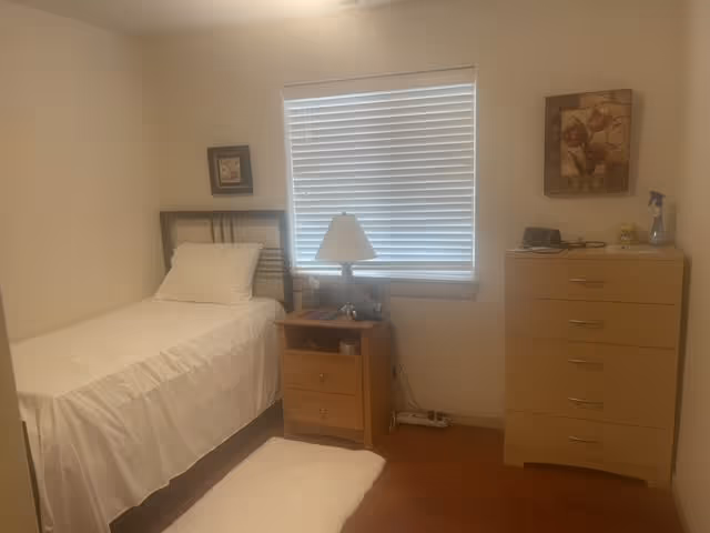 A small bedroom with a single bed covered in white bedding, a wooden nightstand with a lamp and some items on it, a window with closed blinds, a wooden dresser with five drawers, and two framed pictures hanging on the walls.
