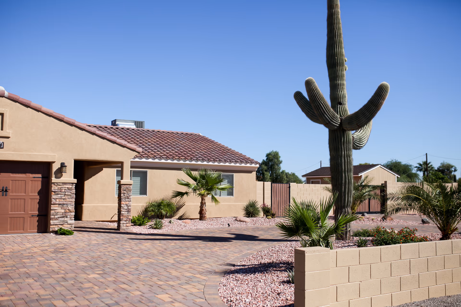Exterior view of a single-story building with a tiled roof and beige stucco walls, surrounded by desert landscaping including a large cactus, small palm trees, and gravel. The driveway is paved with bricks and a low beige block wall is visible in the foreground under a clear blue sky.