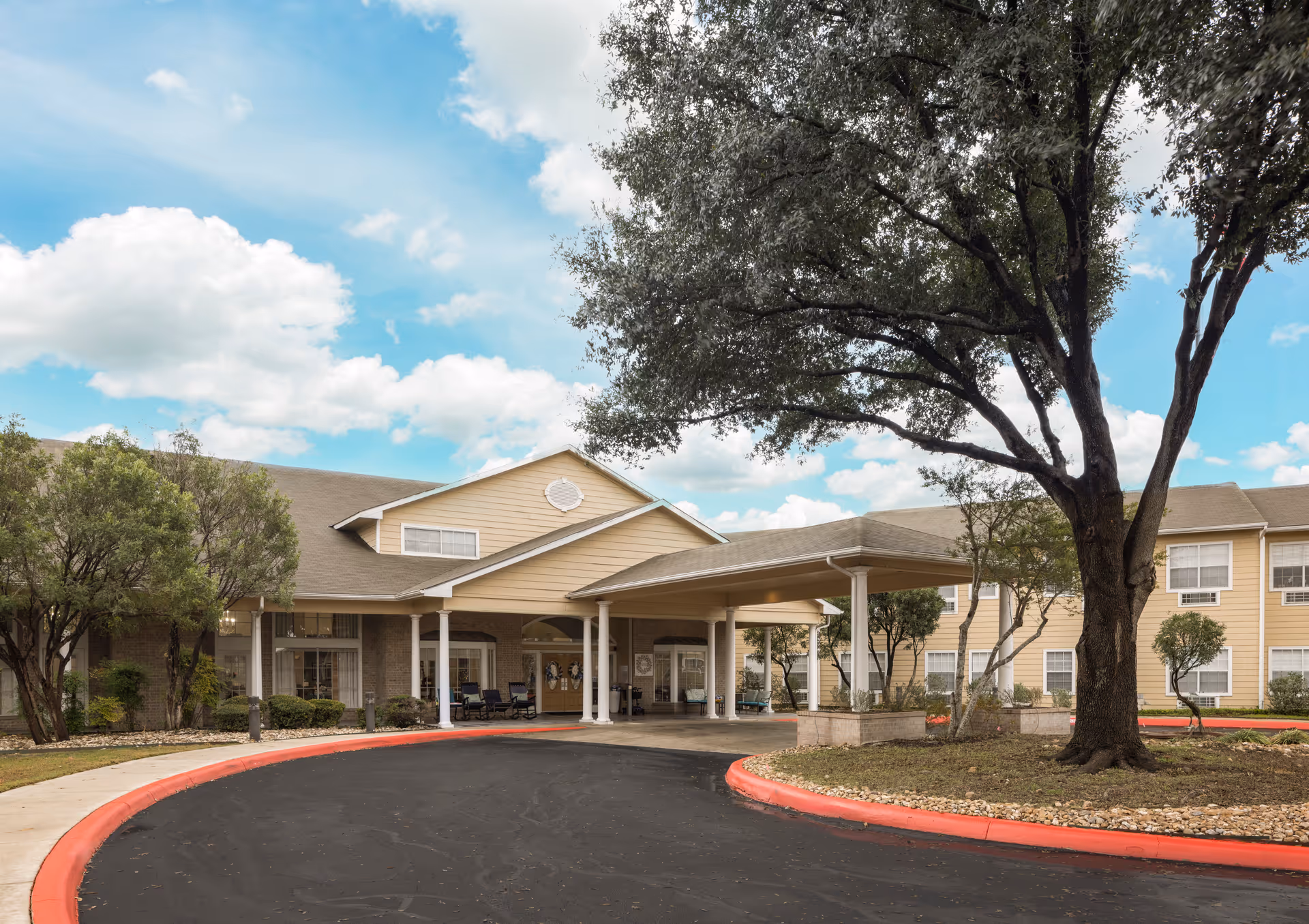 Exterior view of The Waterford at Thousand Oaks senior living facility showing a large covered entrance with white pillars, surrounded by trees and landscaping under a partly cloudy sky.