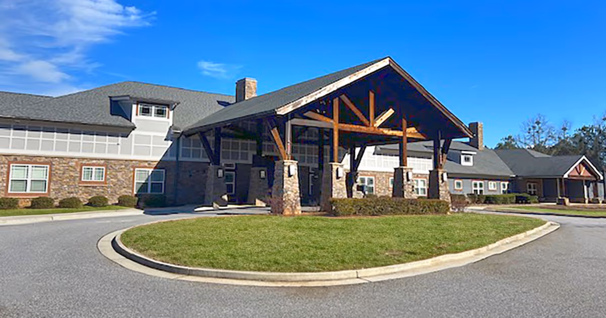 Front exterior view of Legacy Village of Athens, showing a large building with stone and siding facade, a covered entrance supported by wooden beams, a circular driveway, and a well-maintained lawn under a clear blue sky.