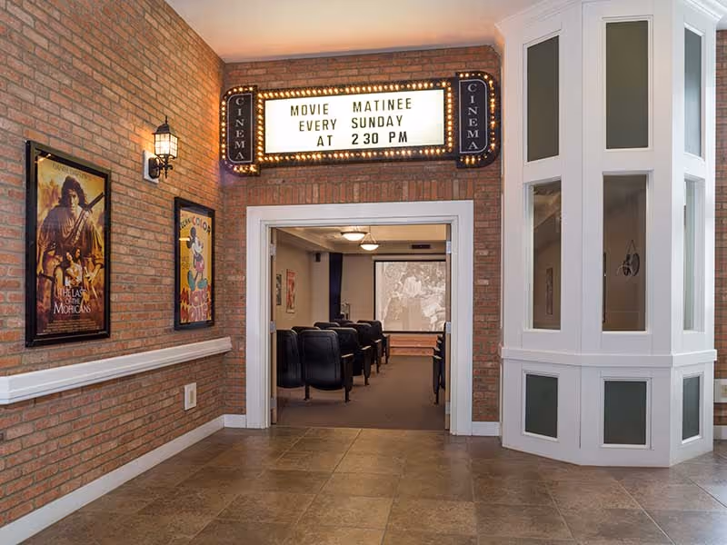 Interior view of a small movie theater room in a senior living facility. The entrance is framed by brick walls with two vintage movie posters on the left side. Above the entrance is a lighted marquee sign that reads 'MOVIE MATINEE EVERY SUNDAY AT 2:30 PM'. Inside the theater room, rows of black cushioned chairs face a screen showing a black and white movie scene. The floor is tiled, and there is a white decorative column structure on the right side.