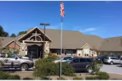 Exterior view of a single-story health care center building with a gabled roof, stone and brick facade, multiple windows, an entrance with a covered porch, several parked cars in front, and an American flag on a flagpole in the foreground under a clear blue sky.