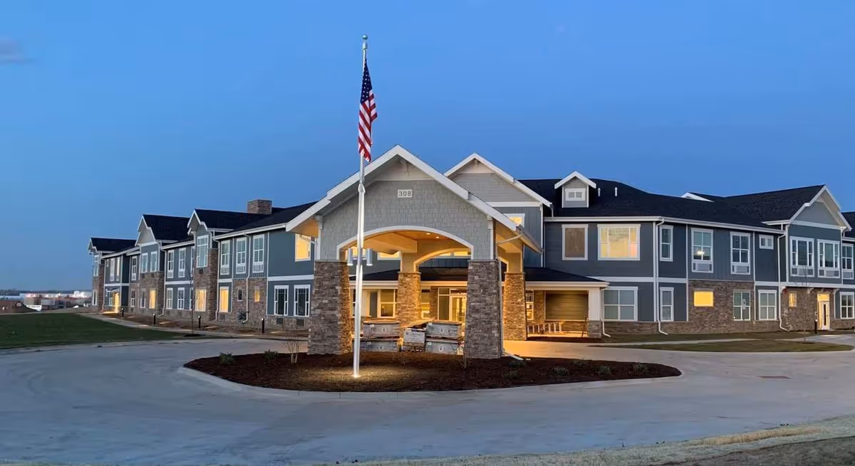 Exterior view of a two-story senior living facility building at dusk with lights on inside. The building has a covered entrance supported by stone pillars and an American flag on a flagpole in front. The sky is clear and blue.