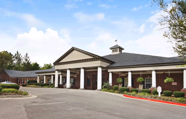 Exterior view of a single-story brick building with white columns supporting a covered entrance. The building has a gray shingled roof with a small cupola on top. There are shrubs and hanging plants along the front, and a paved driveway with parking spaces in front of the entrance. The sky is partly cloudy.