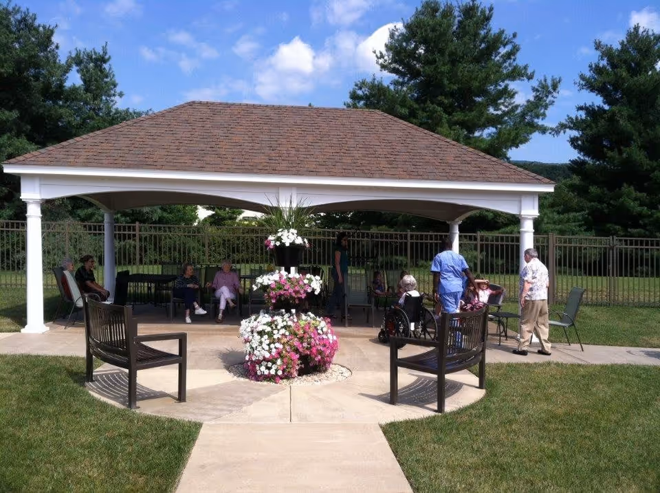 Outdoor gazebo area with a shingled roof and white pillars, surrounded by green grass and trees. Several elderly people are seated on chairs under the gazebo, with some being assisted by caregivers. In the center of the paved area in front of the gazebo is a tiered flower arrangement with pink and white flowers. Two wooden benches face the flower arrangement.