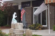 Outdoor view of the entrance area of Dunkirk Rehabilitation and Nursing Center featuring a small statue on a stone pedestal wrapped with rope, an American flag, a white flag on a pole, and a brick building with large windows and a small patio with chairs and a table.